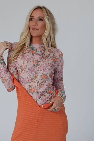 A person with long blonde hair stands against a plain background, wearing the Three Bird Nest Made You Blush Floral Top in Pink Floral that features a fitted long sleeve silhouette and an orange skirt. They accessorize with multiple rings, bracelets, and a necklace. Their gaze is directed to the side.