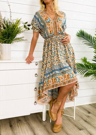 A person wearing the Finn Tie Back High Low Dress from Anna Kaci Wholesale, which features a colorful blue and orange floral print with a cinched waist and high-low hemline, leans against a white dresser. The setting includes indoor plants, wooden floors, and white walls.