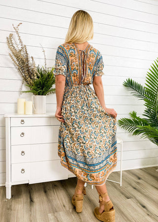 A woman with long blonde hair is standing with her back to the camera, wearing the Finn Tie Back High Low Dress by Anna Kaci Wholesale, which showcases blue and orange bohemian patterns. She stands in a bright room next to a white dresser adorned with plants and candles, with greenery in the background.