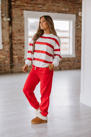 In a well-lit room with exposed brick walls, a woman stands wearing the Callahan Striped Pullover and Joggers Set from Liam & Company. Her outfit includes a white sweatshirt with red stripes and matching red joggers, complemented by tan slippers with white fur trim, hands casually tucked in her pockets.