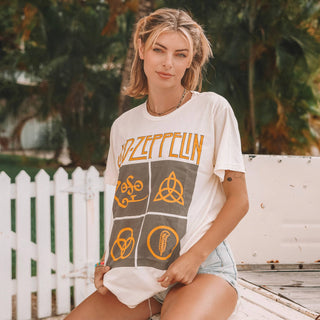 A woman with blonde hair is sitting outdoors near a white picket fence and greenery. She is wearing a white Zoso Led Zeppelin Vintage Tee from Life Clothing Co and knee-length denim shorts. With a relaxed expression, she holds the bottom of her t-shirt.