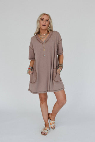 A woman stands against a plain white background, wearing the Brooklyn Soft Touch Tunic Dress from Three Bird Nest. The loose-fitting, boho-chic taupe dress features front pockets. She accessorizes with layered necklaces, bracelets, and rings, and pairs her outfit with patterned sandals. Her blonde hair falls past her shoulders.