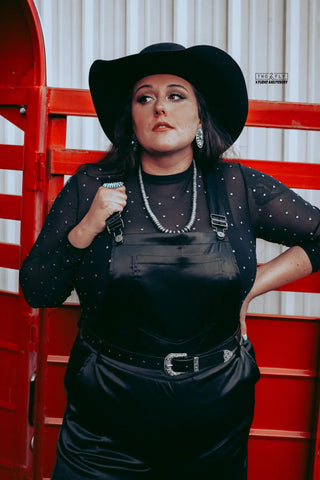 A person dressed in a black hat and a High Rank Black Mesh Top from Western Edge Boutique stands confidently against a red metal structure. They wear silver jewelry, including earrings, a necklace, and a ring, with their right hand raised near their chest.