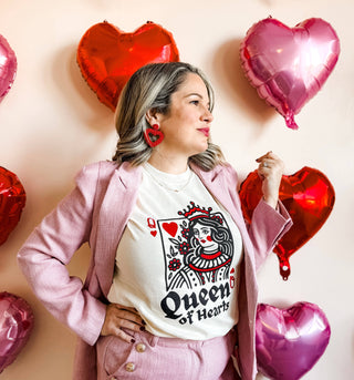 A woman confidently poses in a Mugsby "Queen of Hearts" Valentine Tee paired with a pink blazer, heart-shaped earrings, and surrounded by red and pink heart balloons against a light background.