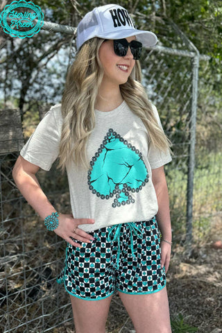 A person stands outdoors wearing a white cap with the word "HOWDY" in black letters, a Sterling Kreek My Lucky Day Tee featuring a large marble turquoise spade illustration, and black-and-white checkered shorts with teal trim. They have long blonde hair, black sunglasses, and a teal bracelet adorned with concho studs.