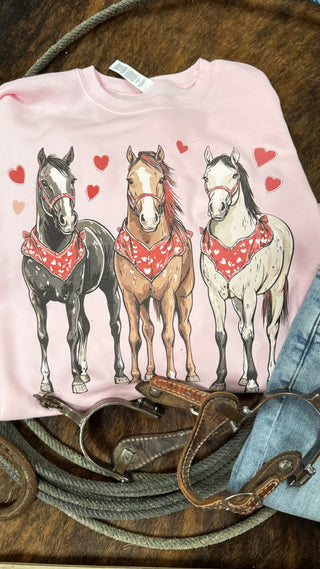 The Giddy Up Valentine from Western Edge Boutique is a pink shirt showing three horses with red bandanas and surrounded by hearts, ideal for horse lovers. Part of a unique collection, it’s styled with coiled rope and leather straps on a textured backdrop.