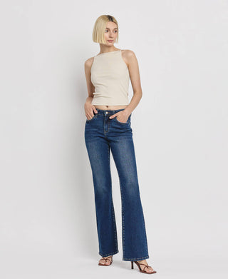 A person with short blonde hair, wearing a beige sleeveless top and Kip Mid Rise Relaxed Bootcut Jeans from Lovervet by VERVET, posing with one hand in their pocket and the other resting on their hip. They are standing against a plain white background, wearing brown heeled sandals.