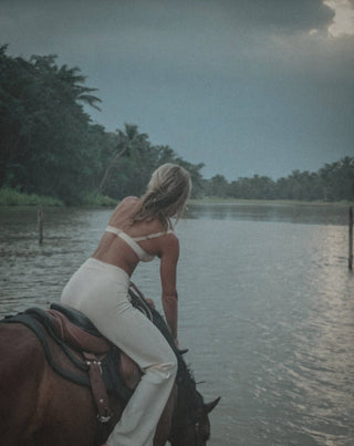 A woman dressed in a white outfit rides a horse through a shallow river under an overcast sky, with lush green trees lining the riverbank in the background. The tranquil, natural atmosphere of the scene evokes the serene fragrance of R. Rebellion's Coastal Cowgirl Candle 8 oz., with its subtle musk notes.