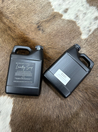 Two black gallon containers of "Laundry is SO much fun when it smells this great!!!" fragrant laundry detergent from Mav Wicks Candle Co are placed on a brown and white fur backdrop. The containers have handles and black labels; one label displays the brand and product details, while the other is partially visible.