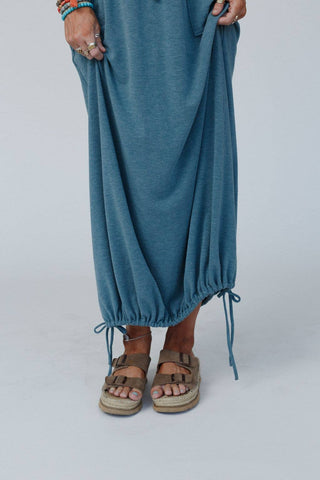 A person is wearing the Rylie Drawstring Maxi Skirt - Teal Boho Skirt from Three Bird Nest, featuring an elastic drawstring waistline and a cinched bottom. They are also sporting brown Birkenstock sandals and several colorful bracelets and rings, with their hands holding up the skirt slightly. The background is plain and light-colored, enhancing their boho western wardrobe.