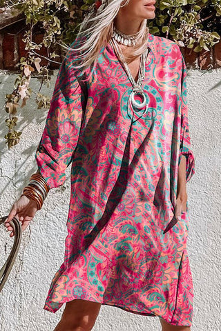 A person stands outdoors wearing the Retro Paisley Tab Sleeve Dress: Rose, a colorful long-sleeve bohemian chic dress by Pretty Bash with pink and green paisley designs. They accessorize with layered necklaces, multiple bangles on one wrist, and their other hand resting in a dress pocket. The backdrop features foliage and a white wall.