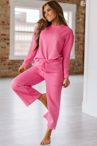 In a bright room with large windows and exposed brick walls, a person playfully poses on one leg wearing the vibrant pink Stockton Textured Set from Liam & Company. The outfit includes a long-sleeve top and pants, each featuring exquisite textures in their subtle pattern.