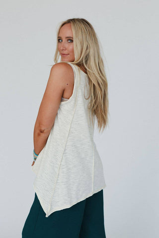 A woman with long blonde hair is facing slightly away from the camera but looking back over her shoulder. She is wearing the Elara Top in neutral by Three Bird Nest and dark-colored pants. The background is plain and neutral.