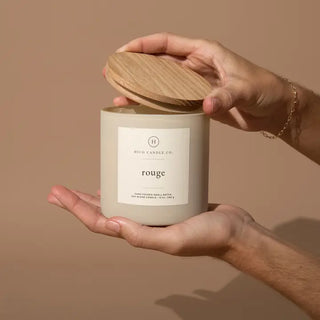 Two hands hold a white candle labeled "Western Edge Boutique - Top Seller Candle," crafted from blended soy wax with a baccarat-inspired aroma. The candle features a wooden lid and is set against a neutral beige background, while the person's arm is adorned with a thin bracelet.