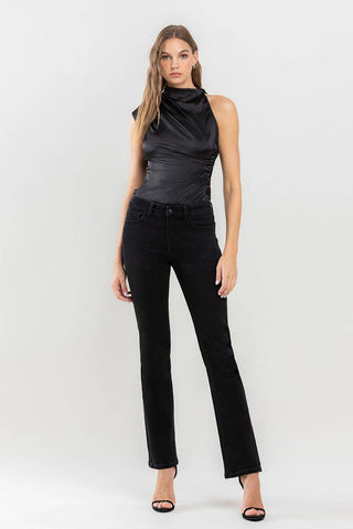 A woman stands against a plain white background, confidently showcasing her wardrobe must-have: a sleeveless, black fitted top paired with Halston Mid Rise Bootcut Jeans from Lovervet by VERVET and open-toe heels. Her long hair is styled in loose waves, highlighting her poised expression.