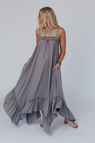 A woman with long blonde hair is wearing the Heavenly Glow Maxi Dress - Mushroom by Three Bird Nest, a flowing grey maxi dress with spaghetti straps and ruffled accents at the bottom. She is smiling and looking to her left, accessorized with layered necklaces and bracelets, radiating boho style in light-colored sandals.