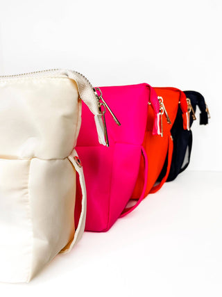 Four Mugsby handbags in a row against a white background, in cream, hot pink, orange, and black. Each bag showcases gold hardware with a sleek design. Crafted from nylon with an outer pocket for convenience.