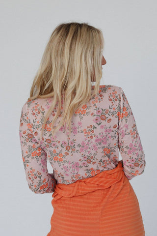 A woman with long blonde hair is facing away from the camera, dressed in the Made You Blush Floral Top - Pink Floral by Three Bird Nest. The semi-sheer, boho blouse features pink, orange, and green flowers and she pairs it with a high-waisted, orange pleated skirt. The background is a plain, light-colored backdrop.