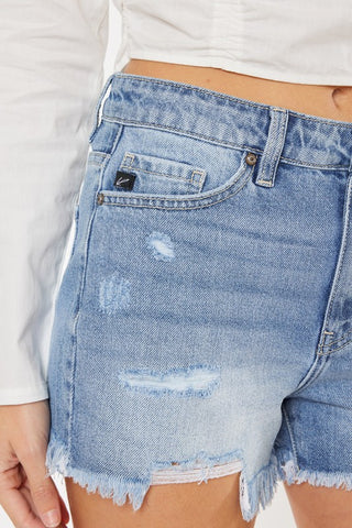 A close-up image showcases a person wearing the Reece Distressed Shorts from VERVET by Flying Monkey. The light blue, high-waisted denim shorts feature frayed hems and distressed details. The person pairs them with a white, long-sleeve top, highlighting only their midsection and upper thigh. A small black tag near the front pocket adds a subtle accent to the comfort denim shorts.