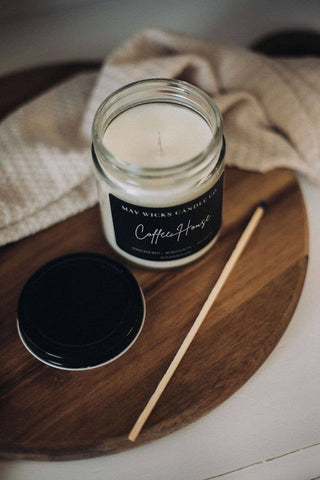 A Luxe(Diva) Candle: 8 OZ in a glass jar with a black label from Mav Wicks Candle Co sits on a wooden surface, making it perfect for home decor. This white candle, featuring one of their best-selling fragrances, has its black lid placed to the side and a thin wooden stick lying next to it. A beige cloth is partially visible in the background.