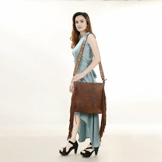 A woman in a light blue, asymmetrical dress and black high-heeled shoes stands against a plain white background. She is carrying the American Darling Large Crossbody Simple Leather with Fringe bag in brown over her shoulder, adding a bohemian flair to her look. Her long hair is down, and she gazes directly at the camera.