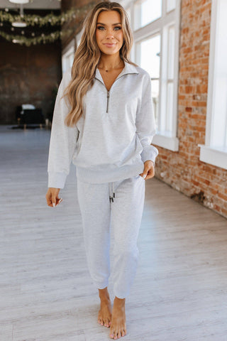 A woman with long, wavy hair is smiling and standing in a room with large windows and brick walls. She is wearing a cozy two-piece Alison Half Zip Loungewear Set from Liam & Company in light gray. The relaxed fit of the half-zip sweatshirt and jogger pants complements her barefoot stance, with her hands relaxed by her sides.