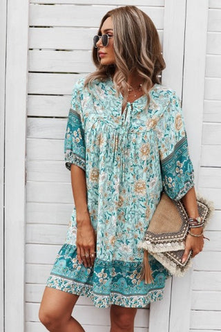 A woman in sunglasses stands against a white wooden wall, wearing the trendy Ruston Floral Boho Dress from Western Edge Boutique in floral turquoise and beige. She holds a woven clutch with tassels, her wavy hair flowing as she looks off to the side on this perfect summer day.