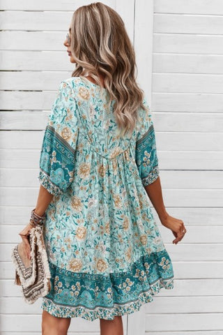 A woman with long, wavy hair is standing against a white wooden wall, wearing the trendy Ruston Floral Boho Dress by Western Edge Boutique in light blue and green. She is holding a textured handbag and looking to the side, showcasing the back of the dress on this summer day.