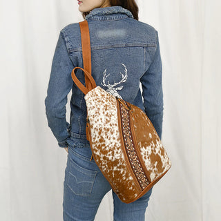 A person wearing a blue denim jacket with an embroidered white design on the back and blue jeans stands with their back facing the camera. They carry an American Darling Collins Brown Cowhide Leather Sling Handbag, featuring intricate patterns on the back zipper area.