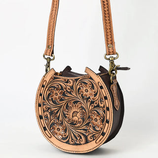 The Bliss Horseshoe Tooled Canteen Purse by American Darling is a circular, tan genuine leather bag featuring intricate floral and vine embossing on the front. It has dark brown sides, a detailed matching strap, brass hardware, and a zipper at the top. The purse is set against a plain white backdrop.