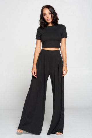 A woman in a stylish Laurel Crop Top and Palazzo Pants Set by Faith Apparel, MADE IN USA, poses confidently. She wears a short-sleeve black crop top paired with flowing, high-waisted wide-leg palazzo pants and light-colored high heels. The background is a simple, light-colored studio setting.