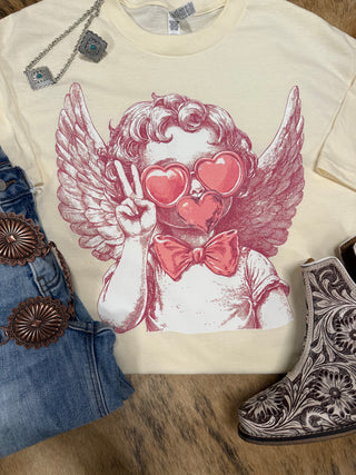 The cream graph tee from Western Edge Boutique's "Cupid Kisses Valentine’s" line features a winged cherub with heart-shaped sunglasses and a bow tie, striking a peace sign. Perfectly paired with denim jeans, a decorative belt, ornamental boots, and jewelry on a textured surface.