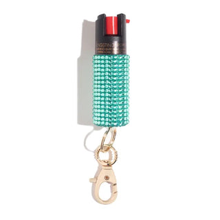 The BLINGSTING Emergency Pepper Spray Key Ring features a teal-colored, rhinestone-studded canister with a black nozzle and red trigger. It includes UV marking dye for added security and a gold keychain clip for easy attachment to bags or key rings. Pictured standing upright against a white background, this product is ideal for women's safety.