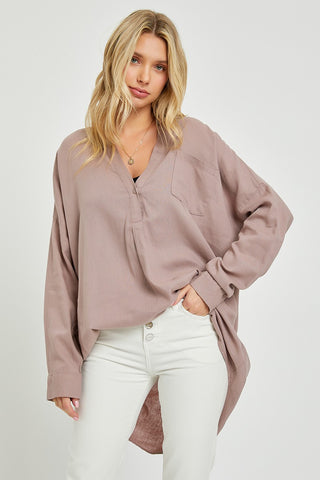 A woman with blonde hair wears a comfortable and stylish Prescott Oversized Popover Linen Shirt by Risen, characterized by its loose-fitting, long-sleeve design in light mauve. She pairs it with a black top underneath and light-colored pants. She stands against a plain white background with a relaxed expression and posture.