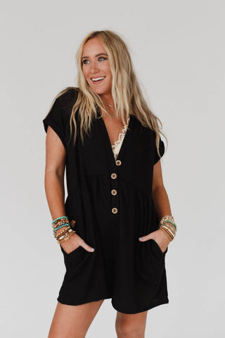 A smiling woman with long blonde hair is wearing the "Sage and Wildflowers Romper" in black by Three Bird Nest. The short-sleeved romper features a deep V-neckline and wooden buttons, giving her a boho-chic vibe. She is adorned with several bracelets on both wrists and has her hands casually tucked into the romper’s pockets, all set against a plain white background.