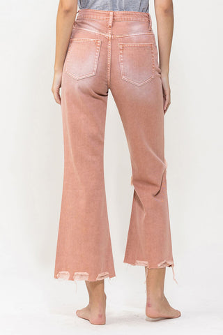 A person is shown from the back, wearing VERVET by Flying Monkey Cove Melon Distressed Vintage Crop Flare Jeans. These light pink, high-waisted jeans feature flared legs with frayed hems and visible front and back pockets, completing the vintage denim look with their distressed edges. The individual is standing barefoot on a neutral background, with the top of a grey shirt also visible.