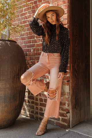 A woman stands outdoors against a brick wall, wearing a wide-brimmed hat, a black floral blouse, and the Cove Melon Distressed Vintage Crop Flare Jeans by VERVET by Flying Monkey. She has one leg raised with her foot resting against the wall and is looking at the camera with a relaxed expression.