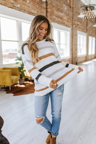 A woman stands in a bright, spacious room with large windows and stylish decor. She is wearing a cozy Mckinley Striped Popcorn Sweater by Liam & Company and ripped jeans, smiling while holding the hem of her sweater. The space has a chic, modern aesthetic with a chandelier and yellow armchair that complements her cozy fashion perfectly.