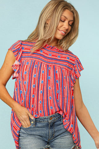 A woman with long blonde hair is smiling and looking down. She is wearing the Kaia Curvy Boho Stripe Mock Neck Top from Western Edge Boutique, which features a red, blue, and white striped pattern. She pairs it with denim shorts and has her right hand in her pocket. The background is a solid light blue.