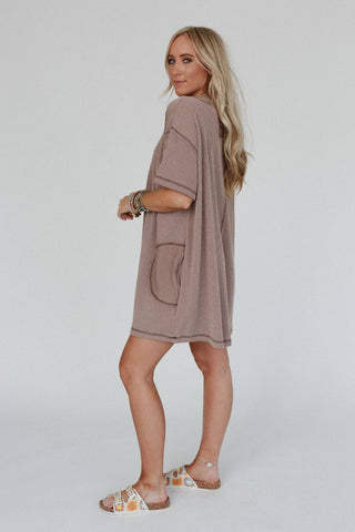 A woman with long blonde hair is wearing an oversized, textured beige Brooklyn Soft Touch Tunic Dress from Three Bird Nest. She pairs it stylishly with white decorated sandals and several bracelets. The background is plain white, and she looks back over her shoulder, exuding effortless charm.