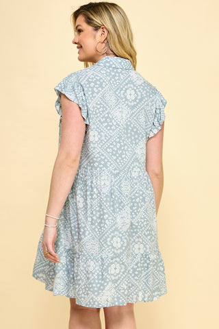 A woman with light blonde hair is shown from behind, wearing the Kota Curvy Tiered Babydoll Dress by Western Edge Boutique. The light blue dress features a white floral pattern, flutter sleeves, a ruffled collar, and a layered ruffle hem. She is standing against a plain beige background.