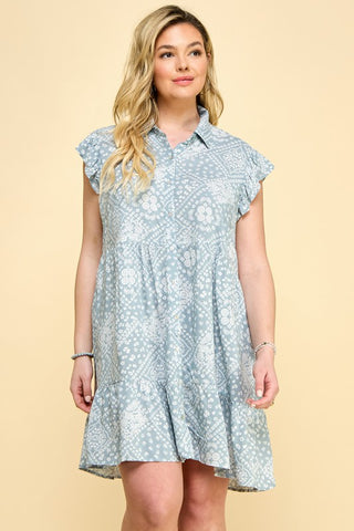 A woman with long blonde hair is standing against a beige background. She is wearing the Kota Curvy Tiered Babydoll Dress from Western Edge Boutique, featuring a light blue pattern, flutter sleeves, and mother of pearl buttons that flares from the waist. Her left arm is adorned with bracelets.