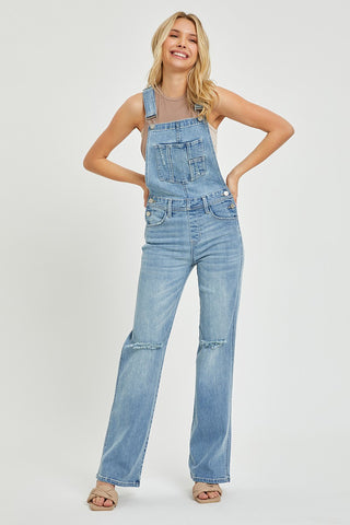 A smiling woman stands with her hands resting on the straps of her light blue, stylish Gannon Curvy Distressed Straight Leg Overalls by Risen. She wears a sleeveless gray top and beige sandals. The background is plain and white.