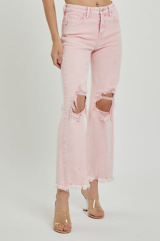 A person is wearing Risen's Memphis Pink Curvy High-Rise Distressed Pants, featuring large rips at the knees and frayed hems. The 98% cotton jeans, with a touch of spandex for stretch, are paired with clear high-heeled sandals. The photo captures them from the waist down against a plain white background.