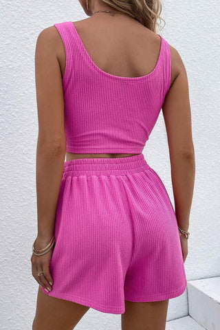 A person with blonde hair is facing away from the camera, wearing the Palmer Unity Rib Knit Crop Tank Top & Shorts Set in bright pink from La Renta Fashion. The background features a white textured wall. The person has bracelets on their right wrist.