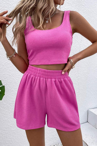 A person with long, wavy hair is wearing the Palmer Unity Rib Knit Crop Tank Top & Shorts Set from La Renta Fashion. The vibrant pink rib knit ensemble features a sleeveless crop tank top and high-waisted shorts with high stretch. They pose with one hand on their hip and the other raised to their hair against a white background.