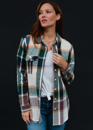 A person with long brown hair is styled in a green, brown, and white flannel shirt from the Panache Accessories collection, specifically the Hunter Plaid Flannel. They wear it over a white top paired with blue jeans and are posing against a black background while holding the shirt with one hand.