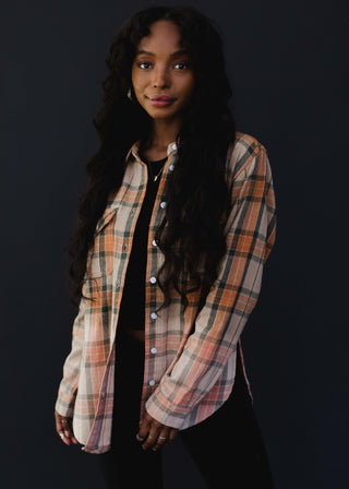 A person with long wavy hair and a slight smile is wearing the Hudson Plaid Flannel by Panache Accessories and black pants, standing against a dark background.