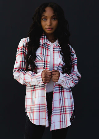 A person dressed in a stylish Sarrow Plaid Flannel from Panache Accessories stands against a black background, wearing it over an elegant ivory top. They have long, wavy hair and are gazing directly at the camera.