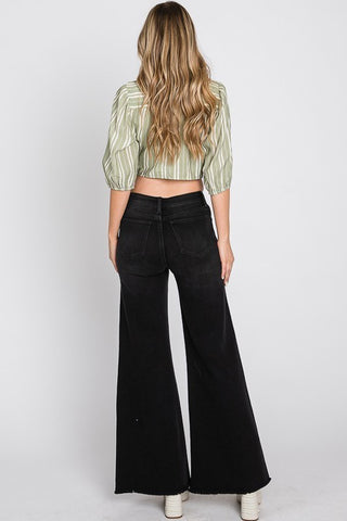 A person with long, wavy hair is standing and facing away from the camera. They are wearing a green and white striped, cropped blouse with puffy sleeves and a pair of HAILEY HIGH RISE BLACK WASHED WIDE LEG FLARE JEANS by Petra153. The background is plain white.